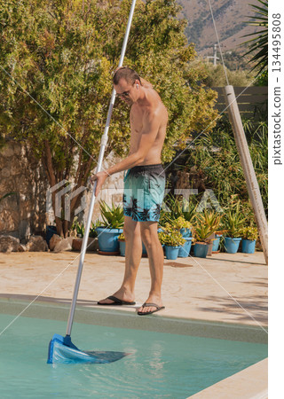 Man in swim trunks with a pool skimmer net cleaning a private swimming pool on a sunny day in a tropical or Mediterranean villa setting 134495808