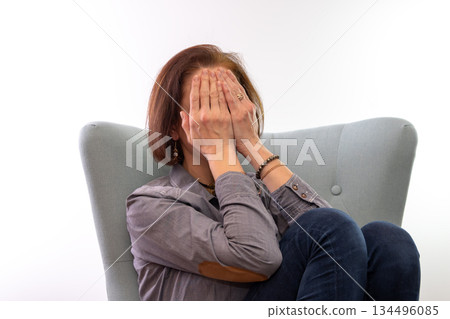 A woman sits in a chair and covers her face with her hands. 134496085