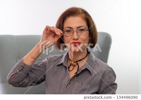 A woman wearing glasses sits in a soft armchair and looks at the camera. 134496090