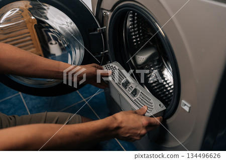 Close-up of person inspecting and removing lint filter from front-loading washing machine or tumble dryer, ensuring proper cleaning and essential appliance maintenance for optimal performance. 134496626
