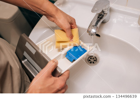 Close-up cropped shot of man cleaning washing machine detergent drawer with sponge at home, performing household chores and maintenance tasks, ensuring appliance hygiene in bathroom laundry area. Close-up cropped shot of man cleaning washing machine detergent drawer with sponge at home, performing household chores and maintenance tasks, ensuring appliance hygiene in bathroom laundry area. 134496639