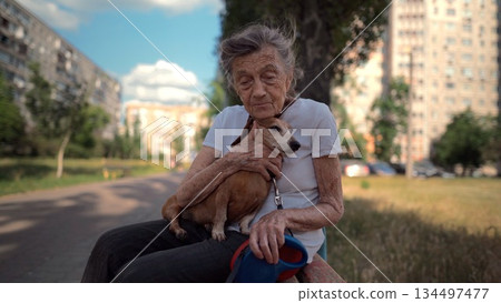 Senior 90-year-old woman with gray hair and deep wrinkles sits outdoors In assisted living facility on bench with small dachshund dog. Old female hugs and cuddles pet in the park on a bench 134497477