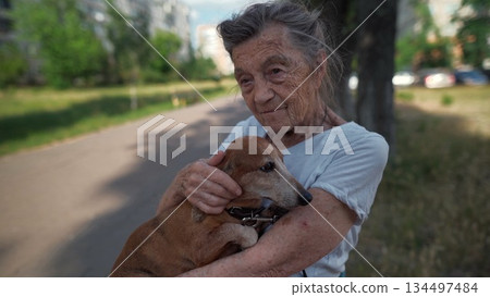 Senior 90-year-old woman with gray hair and deep wrinkles sits outdoors In assisted living facility on bench with small dachshund dog. Old female hugs and cuddles pet in the park on a bench Senior 90-year-old woman with gray hair and deep wrinkles sits outdoors In assisted living facility on bench with small dachshund dog. Old female hugs and cuddles pet in the park on a bench 134497484