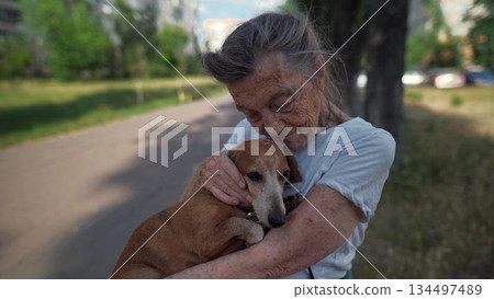 Senior 90-year-old woman with gray hair and deep wrinkles sits outdoors In assisted living facility on bench with small dachshund dog. Old female hugs and cuddles pet in the park on a bench 134497489