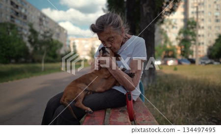 Senior 90-year-old woman with gray hair and deep wrinkles sits outdoors In assisted living facility on bench with small dachshund dog. Old female hugs and cuddles pet in the park on a bench 134497495