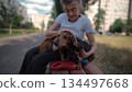 Senior 90-year-old woman with gray hair and deep wrinkles sits outdoors In assisted living facility on bench with small dachshund dog. Old female hugs and cuddles pet in the park on a bench 134497668