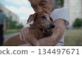 Senior 90-year-old woman with gray hair and deep wrinkles sits outdoors In assisted living facility on bench with small dachshund dog. Old female hugs and cuddles pet in the park on a bench 134497671