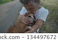 Senior 90-year-old woman with gray hair and deep wrinkles sits outdoors In assisted living facility on bench with small dachshund dog. Old female hugs and cuddles pet in the park on a bench 134497672