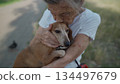 Senior 90-year-old woman with gray hair and deep wrinkles sits outdoors In assisted living facility on bench with small dachshund dog. Old female hugs and cuddles pet in the park on a bench 134497679