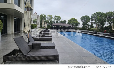 A serene poolside with wicker loungers, closed umbrellas A serene poolside with wicker loungers, closed umbrellas 134497786