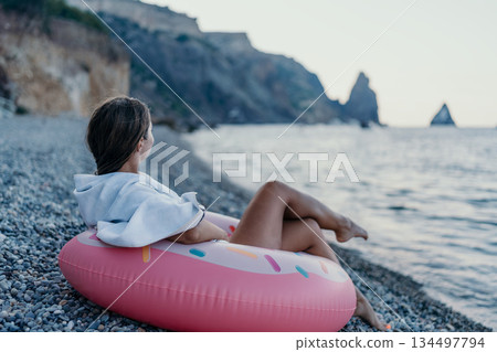 Woman, beach, ocean. Young woman relaxes on pebble beach with donut float, gazing at calm ocean, perfect for tranquil summer travel. Woman, beach, ocean. Young woman relaxes on pebble beach with donut float, gazing at calm ocean, perfect for tranquil summer travel. 134497794