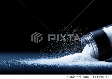 Spilled salt on black surface with grains scattering in dramatic lighting creating sense of motion and contrast perfect for culinary or kitchen themes Spilled salt on black surface with grains scattering in dramatic lighting creating sense of motion and contrast perfect for culinary or kitchen themes 134498916