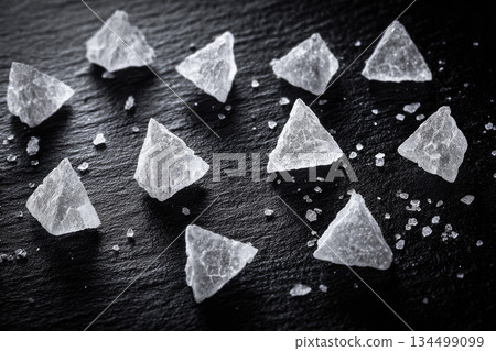 Fleur de sel flakes arranged on dark stone surface create striking visual with their crystalline texture and natural geometric shapes, evoking sense of culinary elegance 134499099