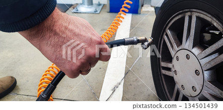A close-up of an adult's hand inflating a car tire with a compressed air hose at a gas station or garage. Concept: vehicle maintenance, car care, road safety. A close-up of an adult's hand inflating a car tire with a compressed air hose at a gas station or garage. Concept: vehicle maintenance, car care, road safety. 134501439
