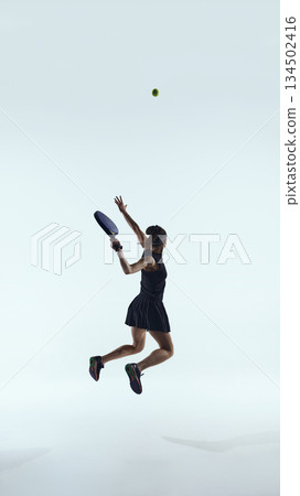 Female padel player jumps mid-air to hit the ball against clean studio background. 134502416