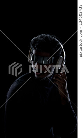 Man wearing futuristic headphones and visor touching earpiece in dark studio Man wearing futuristic headphones and visor touching earpiece in dark studio 134502435