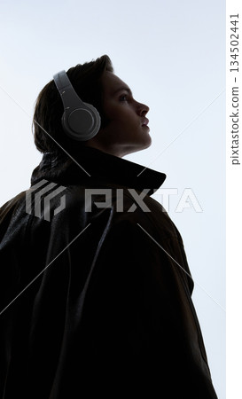 Side view portrait of young man wearing headphones and leather jacket against bright background. Side view portrait of young man wearing headphones and leather jacket against bright background. 134502441