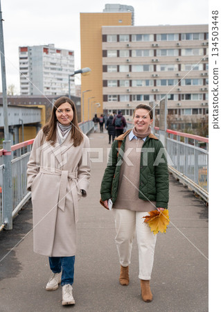 Two women walk together on a pedestrian bridge, smiling and talking, autumn city background, neutral tones, casual outerwear, relaxed mood Two women walk together on a pedestrian bridge, smiling and talking, autumn city background, neutral tones, casual outerwear, relaxed mood 134503448