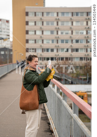 Woman in a green jacket stands on a pedestrian bridge, holding a smartphone and yellow autumn leaves against an urban residential background Woman in a green jacket stands on a pedestrian bridge, holding a smartphone and yellow autumn leaves against an urban residential background 134503449