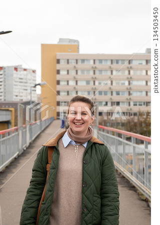 A smiling woman in a green quilted jacket walks on a pedestrian bridge with apartment buildings in the urban background under an overcast sky 134503450