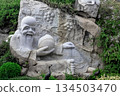 A stone sculpture of a sage against a backdrop of cliffs and greenery at the Heavenly Grottoes Park, Hainan. 134503470