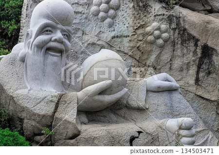 A stone sculpture of a sage against a backdrop of cliffs and greenery at the Heavenly Grottoes Park, Hainan. 134503471