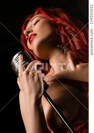 Woman sings into a microphone while performing on stage during a night event Woman sings into a microphone while performing on stage during a night event 134503832