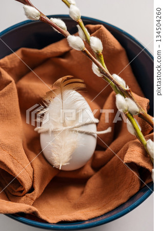 Easter background. Willow branches, white egg with feather in a blue bowl. Spring time. Minimalistic and bright concept Easter background. Willow branches, white egg with feather in a blue bowl. Spring time. Minimalistic and bright concept 134504260