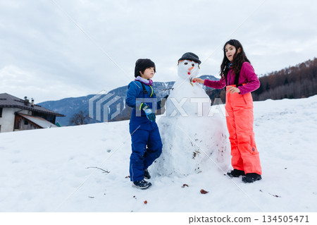 two children making snowman against mountain background 134505471