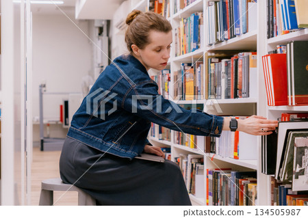 woman is sitting at shelves in library, student is choosing book. library is filled with books. 134505987