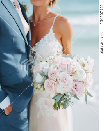 bridal bouquet of pale pink roses and white flowers held by a bride in lace dress, with groom in a blue suit beside her, at romantic beach wedding, perfect for romance, floral design, wedding themes. 134507993
