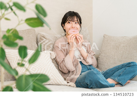 一位年輕的亞裔女性坐在客廳的沙發上吃甜甜圈。 134510723