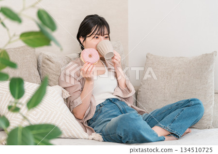 一位年輕的亞裔女性坐在客廳的沙發上吃甜甜圈。 134510725