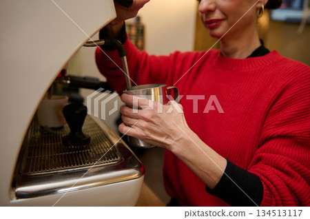 A cafe owner in a red sweater pours milk from a metal pitcher into a steaming espresso machine, conveying hospitality, skill, and daily cafe operation. A cafe owner in a red sweater pours milk from a metal pitcher into a steaming espresso machine, conveying hospitality, skill, and daily cafe operation. 134513117