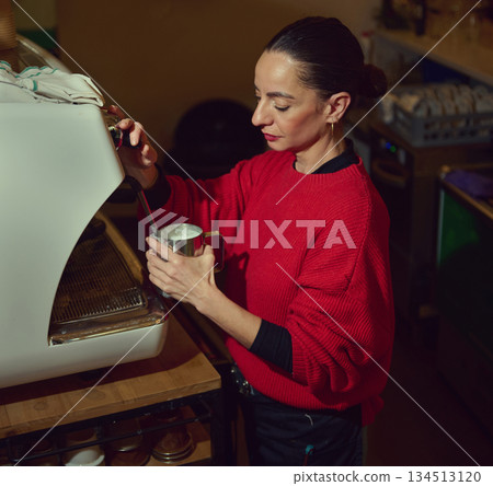 A Hispanic cafe owner in a red sweater brews milk foam at an espresso machine, creating a warm, focused moment of hospitality in a small cafe setting. 134513120