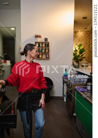 A smiling cafe owner in a red knit sweater and apron greets guests near the counter in a warm, busy cafe with shelves, plants, and dishes. A smiling cafe owner in a red knit sweater and apron greets guests near the counter in a warm, busy cafe with shelves, plants, and dishes. 134513121