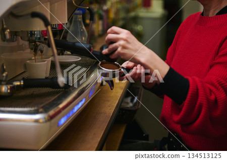 A Hispanic middle-aged cafe worker operates a professional espresso machine, preparing a shot for customers. Warm red sweater, bustling counter, focus on craft, hospitality, and small-business cafe 134513125