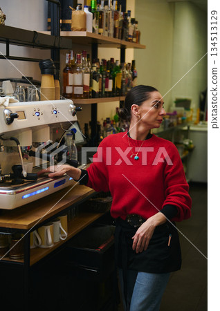 A Hispanic cafe owner in a red sweater works at an espresso machine in a cozy cafe, with shelves of bottles behind her, projecting hospitality and small business vibes. 134513129