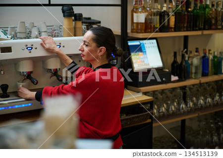 A Hispanic woman runs a cafe, operating an espresso machine and serving customers. The busy bar highlights hospitality, small business energy, and daily cafe routines. 134513139