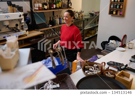 Hispanic cafe owner in a red sweater stands behind a busy bar, smiling as she manages drinks and dishes in a cozy cafe setting. Hispanic cafe owner in a red sweater stands behind a busy bar, smiling as she manages drinks and dishes in a cozy cafe setting. 134513146