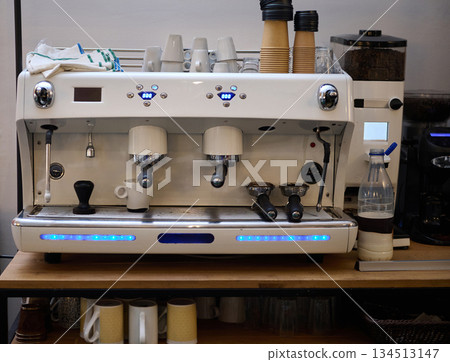 A sleek white espresso machine sits on a cafe counter with stacked cups, a grinder, and a bottle of water nearby. Bright blue LEDs highlight the modern cafe setup. A sleek white espresso machine sits on a cafe counter with stacked cups, a grinder, and a bottle of water nearby. Bright blue LEDs highlight the modern cafe setup. 134513147