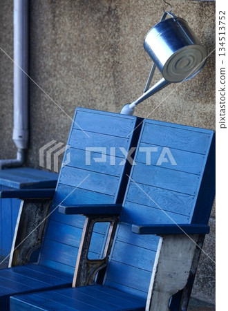 A blue chair and a silver watering can placed along the wall of a building on the street A blue chair and a silver watering can placed along the wall of a building on the street 134513752