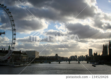 view of the London Eye on the River Thames under a clear blue sky, with clouds and a city skyline along the riverbank, suggesting travel, tourism and urban imagery 134520426