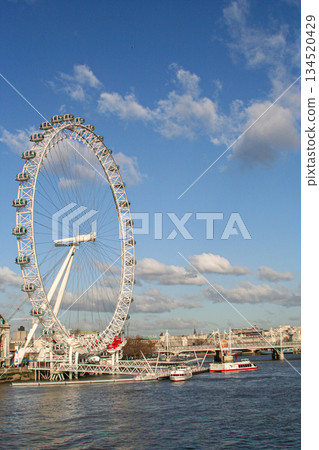 view of the London Eye on the River Thames under a clear blue sky, with clouds and a city skyline along the riverbank, suggesting travel, tourism and urban imagery 134520429