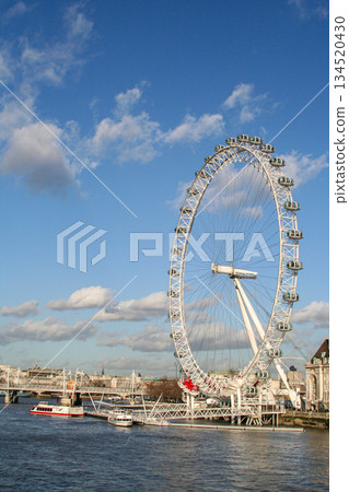 view of the London Eye on the River Thames under a clear blue sky, with clouds and a city skyline along the riverbank, suggesting travel, tourism and urban imagery 134520430