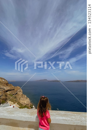 Child enjoying the stunning cliffside view over the water on a sunny day in the picturesque Greek island of Santorini 134523114