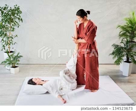 Masseuse making thai yoga massage. Woman therapist doing traditional massage treatment using elbow for female patient. Client lying on mat, meditating, enjoying hand massage in calm atmosphere. Masseuse making thai yoga massage. Woman therapist doing traditional massage treatment using elbow for female patient. Client lying on mat, meditating, enjoying hand massage in calm atmosphere. 134523673