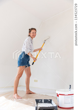 Portrait young adult beautiful single happy woman painting wall paint roller brush white color living room moving new home flat . Female painter decorating interior renovation residential apartment 134523729