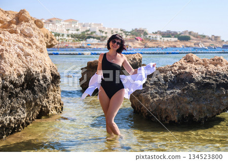 A woman poses on the beach standing in the water near a large rock 134523800