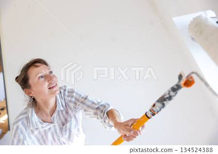 Portrait young adult beautiful single happy woman painting wall paint roller brush white color living room moving new home flat . Female painter decorating interior renovation residential apartment 134524388